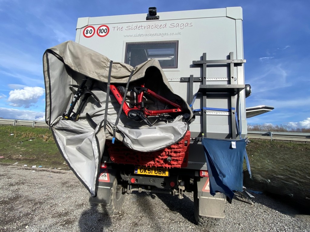 A rear view of a camper van with bikes partially covered by a grey protective cover, showing the branding 'The Sidetracked Sagas' on the back. The setting includes a gravel area with grass and a road in the background under a clear blue sky.