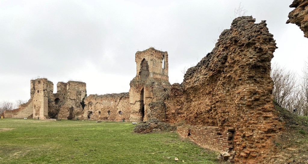 Ruins of an ancient stone wall and towers, set against a cloudy sky with grass in the foreground.