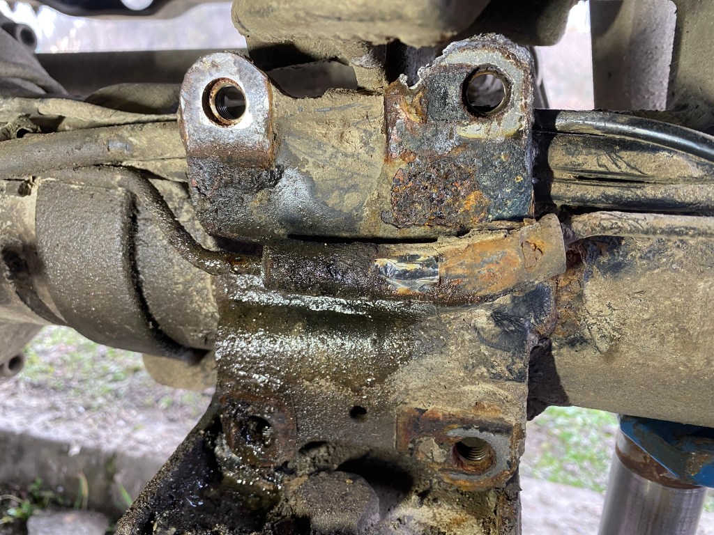 Close-up of a rusty vehicle component showing corrosion and grime, with visible bolts and pipes.