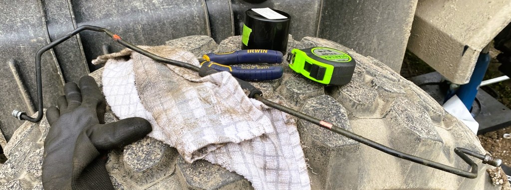 A close-up of a dirty work surface with a black metal rod, a pair of gloves, a measuring tape, and two tools resting on a crumpled cloth, all placed on a large, textured tyre.