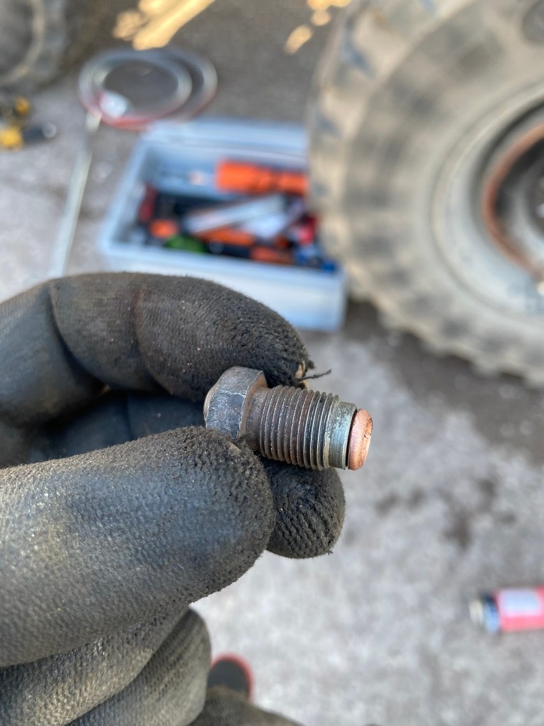 A close-up of a hand wearing a black glove, holding a metal bolt with thread and a copper washer, set against a background of tools and a tyre.