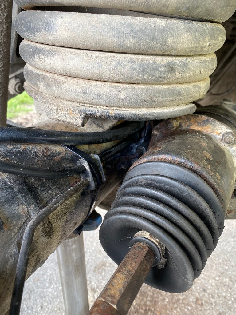 Close-up view of a vehicle's suspension system, showing a coiled rubber spring and a worn rubber boot over a metal rod, with dirt and grime visible.