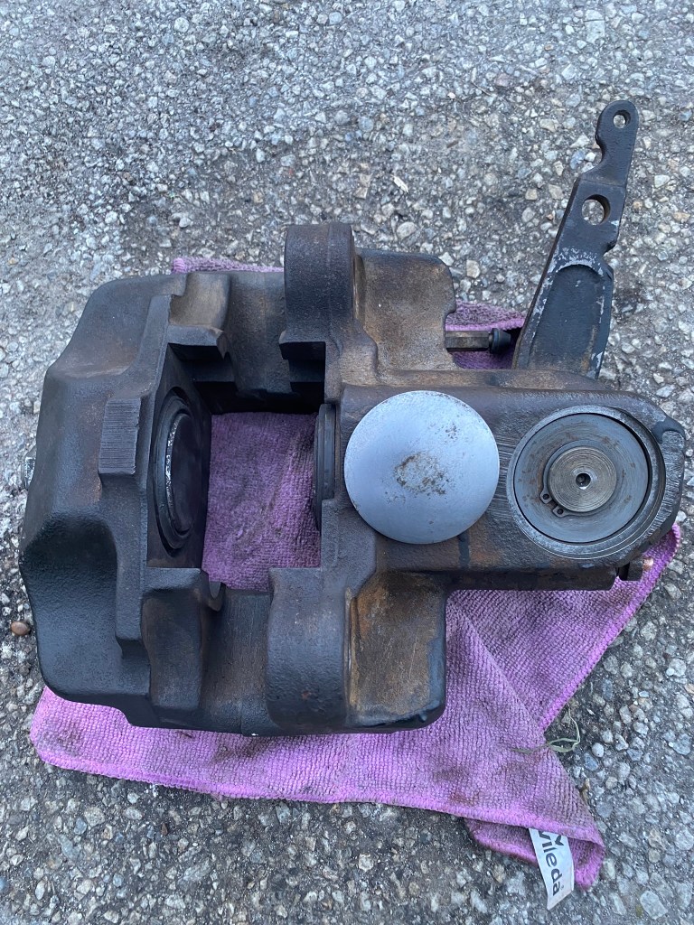 A disassembled brake caliper resting on a pink cloth, displayed on a gravel surface.