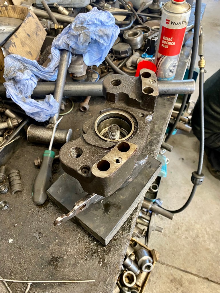 An industrial workbench cluttered with various metal parts and tools, featuring a close-up of a component with multiple holes and a rotating mechanism, alongside a can of cleaner and a blue cloth.