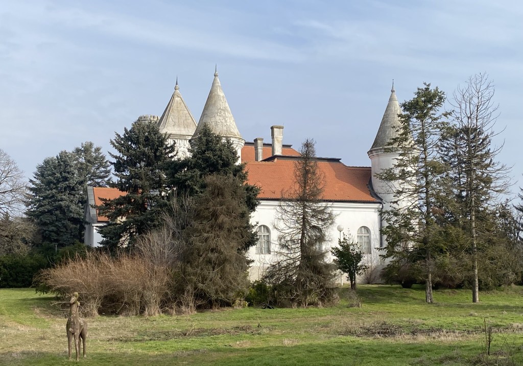 A white castle-like building with grey and pointed towers, surrounded by trees and overgrown vegetation in a grassy area.