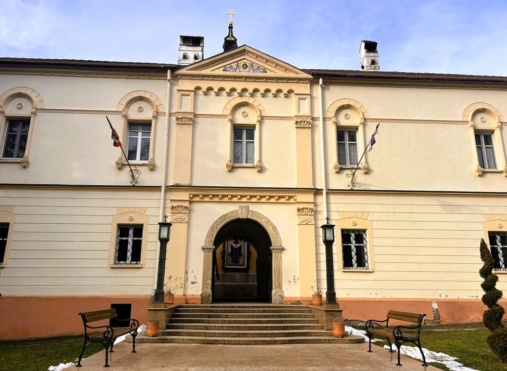 Facade of a historic building with ornate architectural details, featuring an arched entrance, decorative windows, and flags. Benches are positioned on either side of the entrance, with greenery and remnants of snow in the foreground.