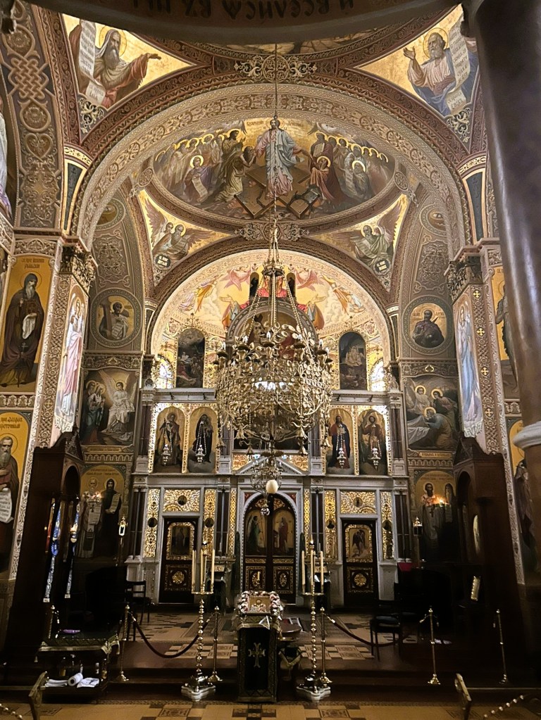 Interior view of an ornate church featuring intricate frescoes and religious icons on the walls, a large chandelier in the centre, and an altar area with candles and decorative elements.