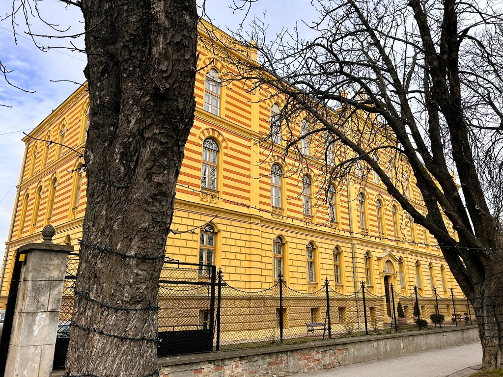 A historic building with yellow and orange striped exterior, viewed from the street, surrounded by bare trees and a metal fence.