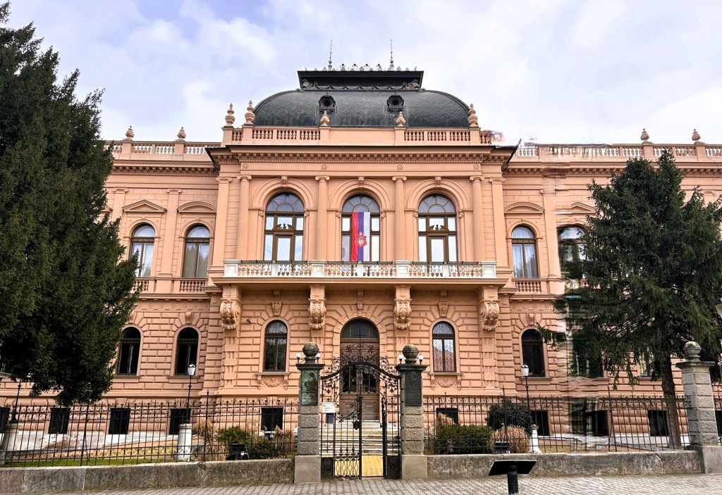 Front view of an ornate pink historical building with large windows and a decorative roof, flanked by tall trees.