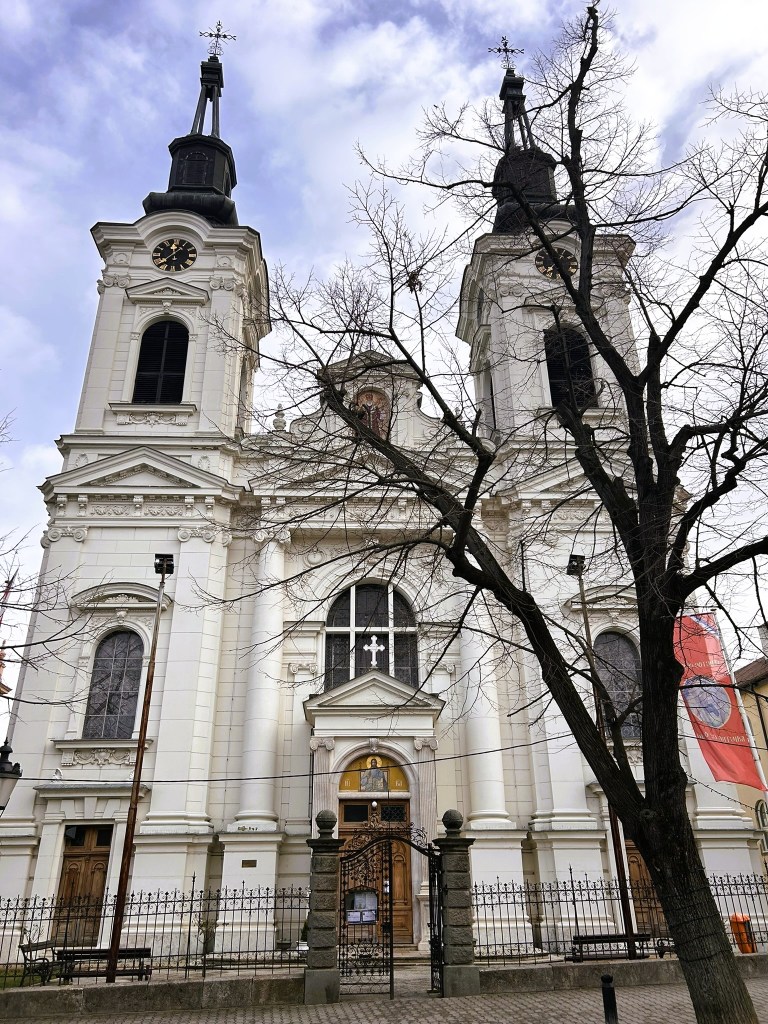 Front view of a grand white church with two tall spires, featuring a clock and decorative architecture, surrounded by leafless trees.
