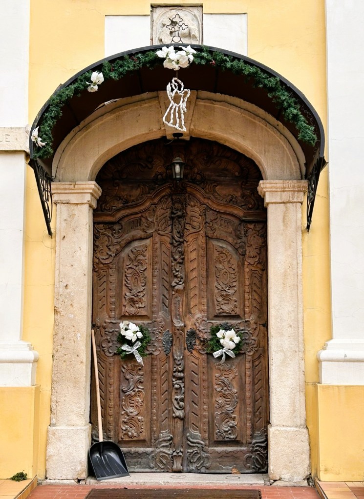 Intricately carved wooden door adorned with white flowers and greenery, beneath a decorative awning featuring an angel figure.