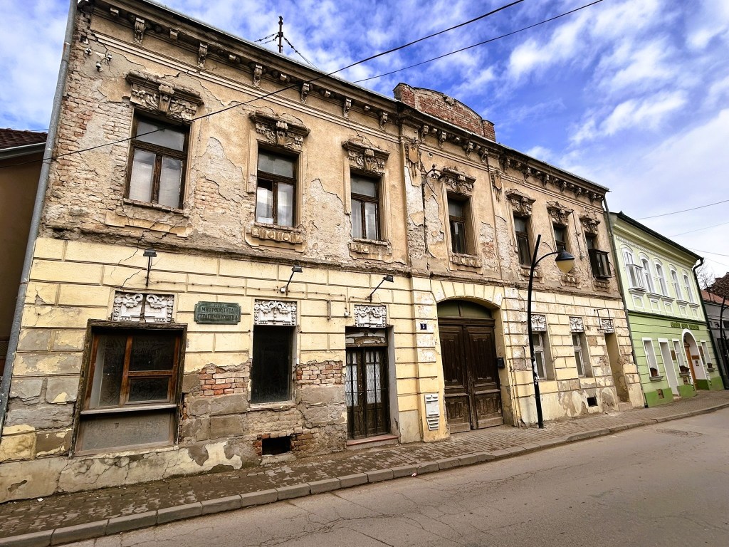 A historic building with a weathered facade, featuring peeling paint and decorative elements above the windows, located on a street with a cobbled pavement.