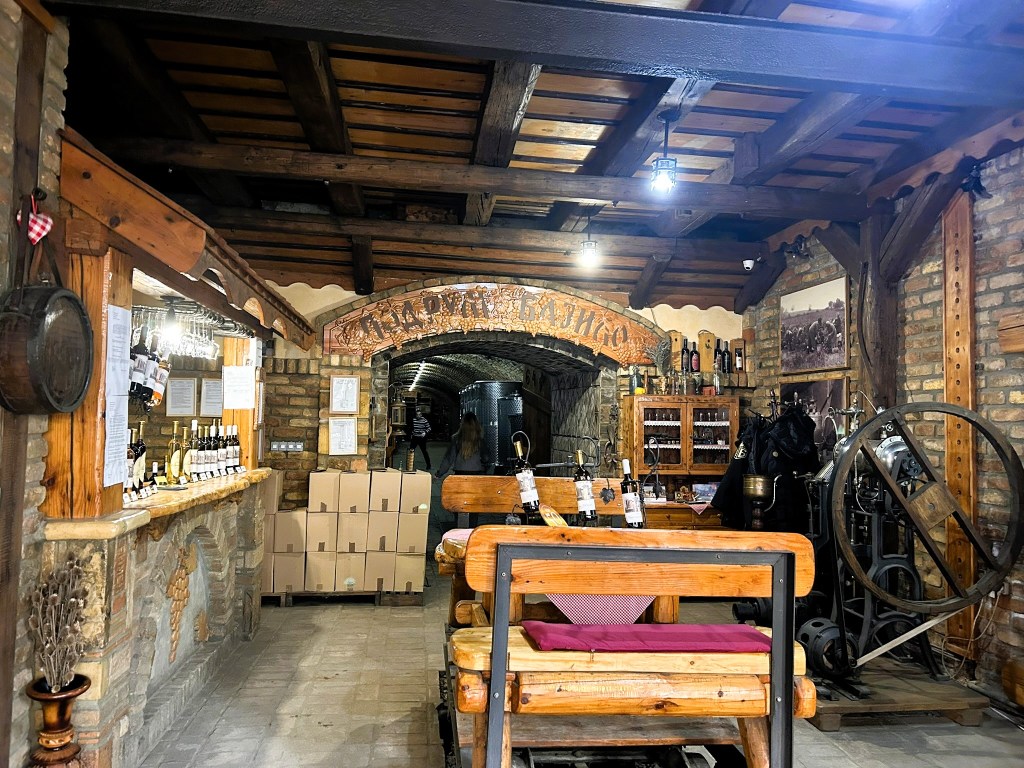 Interior of a rustic wine cellar featuring wooden beams, brick walls, a bar area with wine bottles, and boxes stacked in the background.