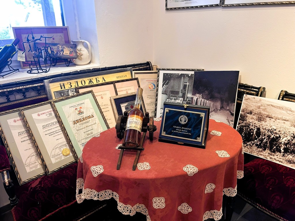 A small round table covered with a lace doily, displaying a bottle of Bermet, several framed diplomas and awards, and black-and-white historical photographs, with a window and decorative items in the background.