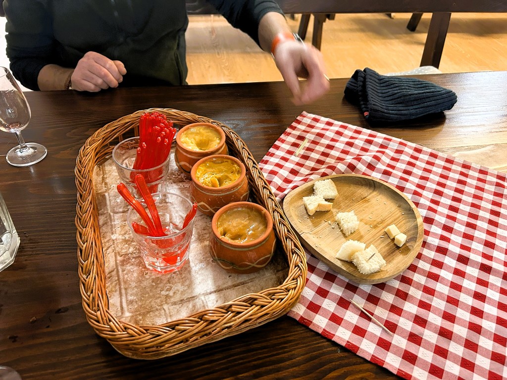 A wooden tray with several clay pots containing sauces, alongside glasses filled with red straws, placed on a checkered tablecloth. A small wooden plate holds pieces of bread and cheese.