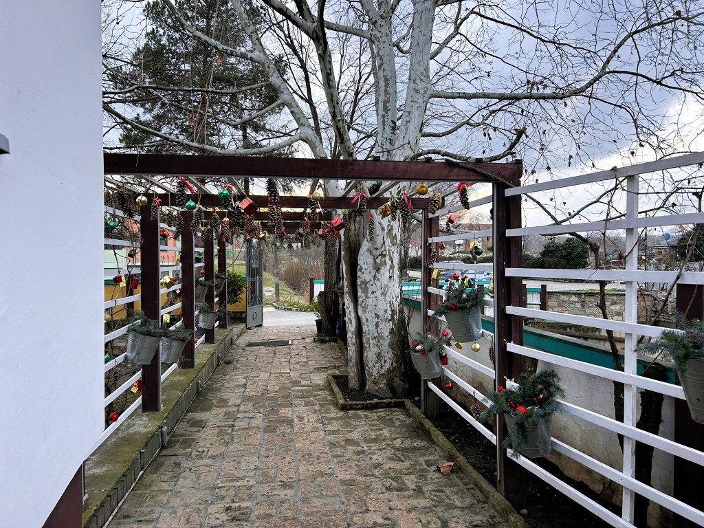 A pathway decorated with festive ornaments and green foliage, flanked by wooden support beams and a large tree, leading into a tranquil outdoor space.