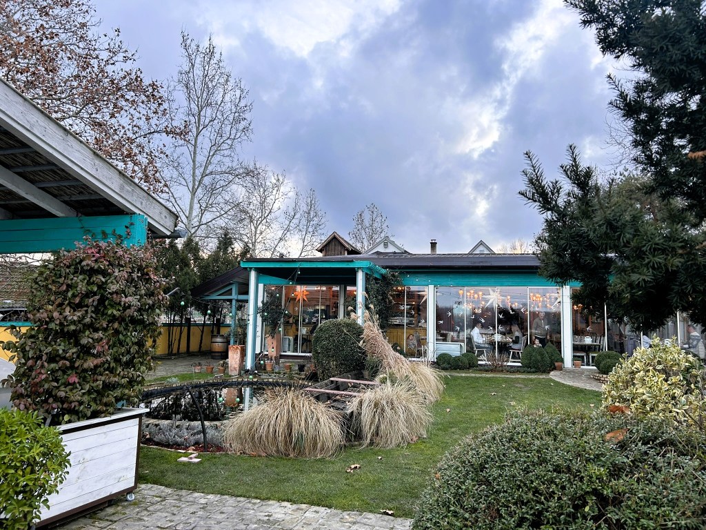 A charming restaurant view featuring large glass windows, surrounded by greenery and a serene garden with ornamental plants and a small pond.