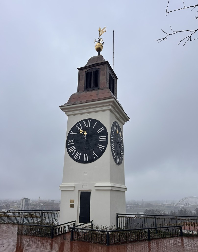 A tall clock tower with a black clock face and Roman numerals, topped with a weather vane, set against a grey, foggy sky.