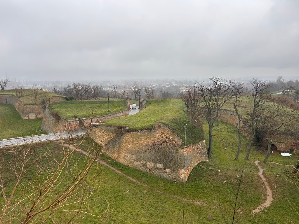 A scenic view of ancient fortification ruins covered in grass, surrounded by barren trees and a cloudy sky.