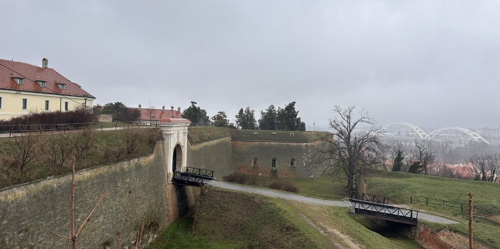 A view of an old fortified structure with a grassy area and a stone pathway, featuring a bridge leading to an archway. The background includes more buildings and a modern bridge under a cloudy sky.