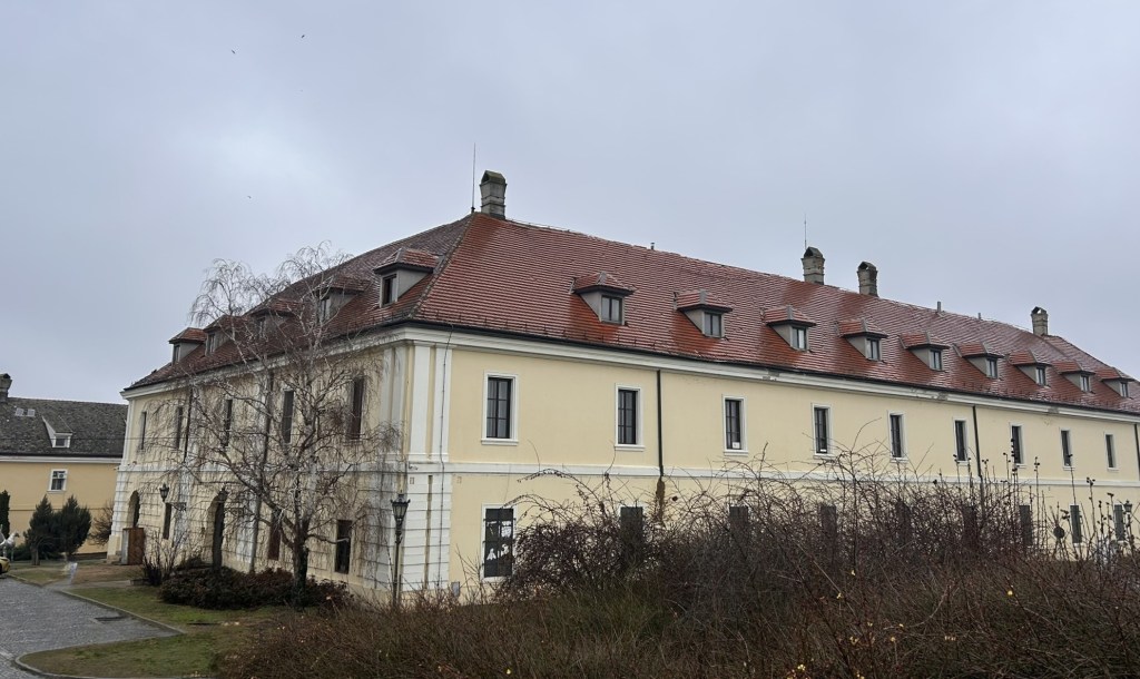 A historical building with a yellow exterior and a red-tiled roof, featuring multiple dormer windows. The structure is surrounded by bare trees and shrubs, under a cloudy sky.