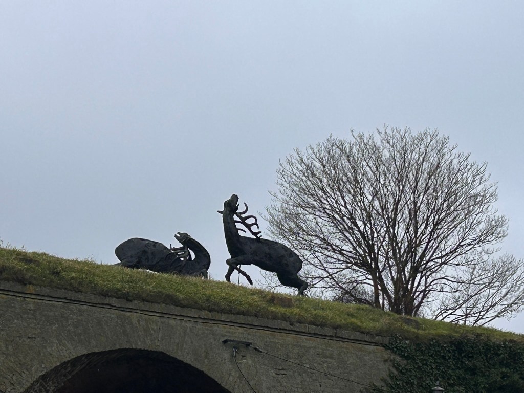 Silhouette of two bronze deer sculptures on a grassy slope, with a bare tree in the background under a cloudy sky.