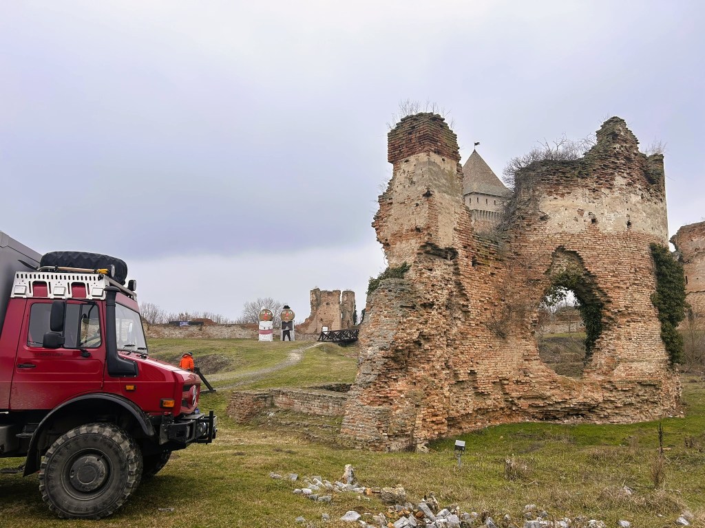 A red all-terrain vehicle parked near crumbling brick ruins of a castle, with a tower in the background and two painted figures standing on a grassy hillside.