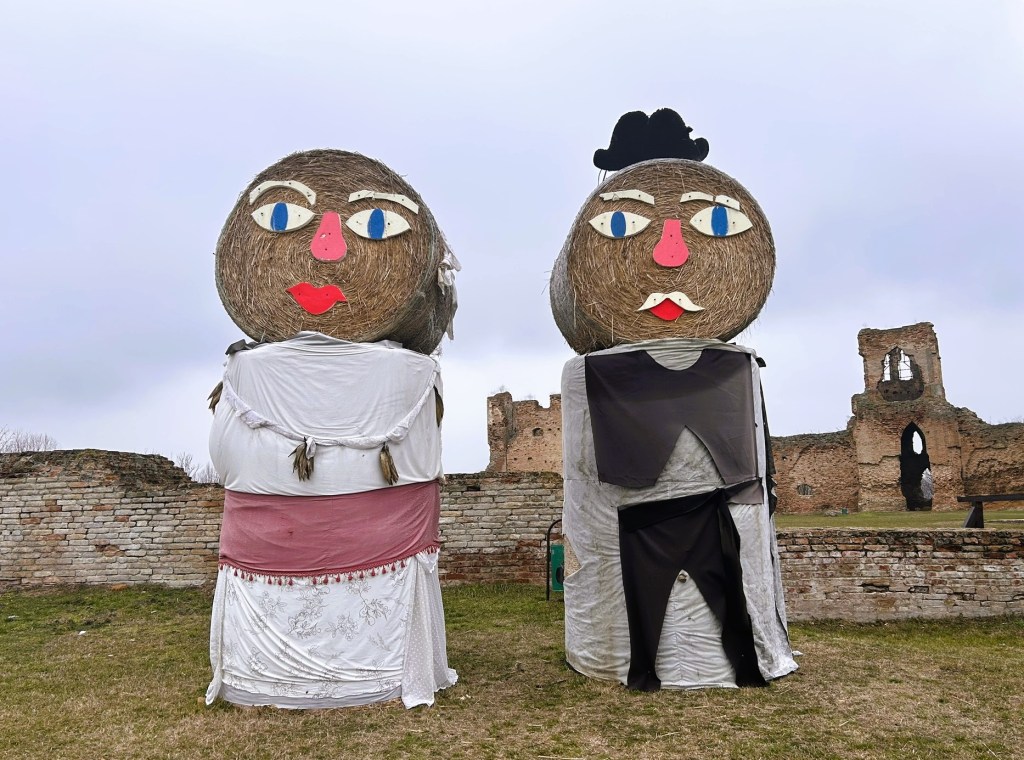 Two large straw figures dressed in clothes, resembling a man and a woman, stand in front of a ruined brick wall under a cloudy sky.