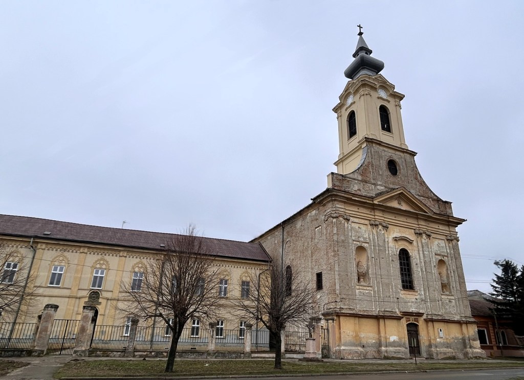 A side view of an old church building with a bell tower and an adjacent historical structure, set against a cloudy sky.