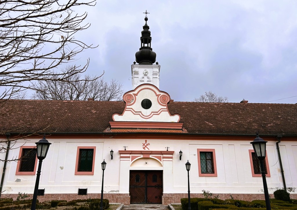 A historic building featuring a pink and white façade with arched windows and a prominent tower topped with a cross, set against a cloudy sky.