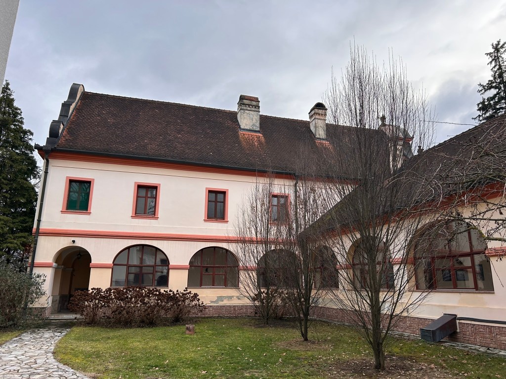View of a historic building with arched windows and a red-tiled roof, surrounded by grassy areas and bare trees.