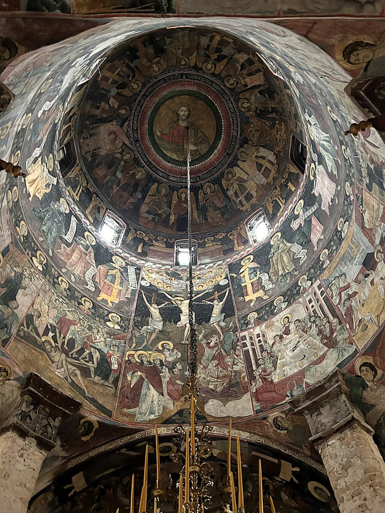 Interior view of a church dome featuring intricate frescoes depicting biblical scenes and figures, with a central image of Christ surrounded by saints and various religious motifs.
