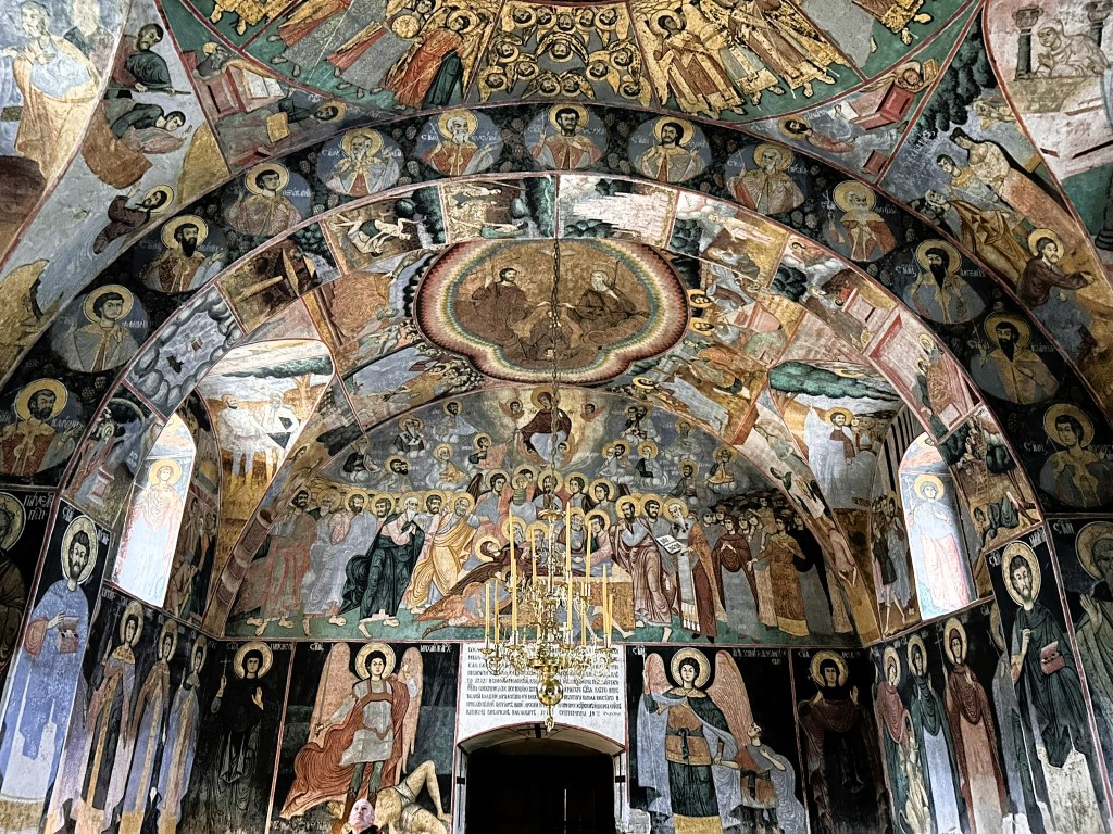 Interior view of a historic church featuring a ceiling adorned with intricate frescoes depicting religious figures, saints, and scenes from the Bible, illuminated by a central chandelier.
