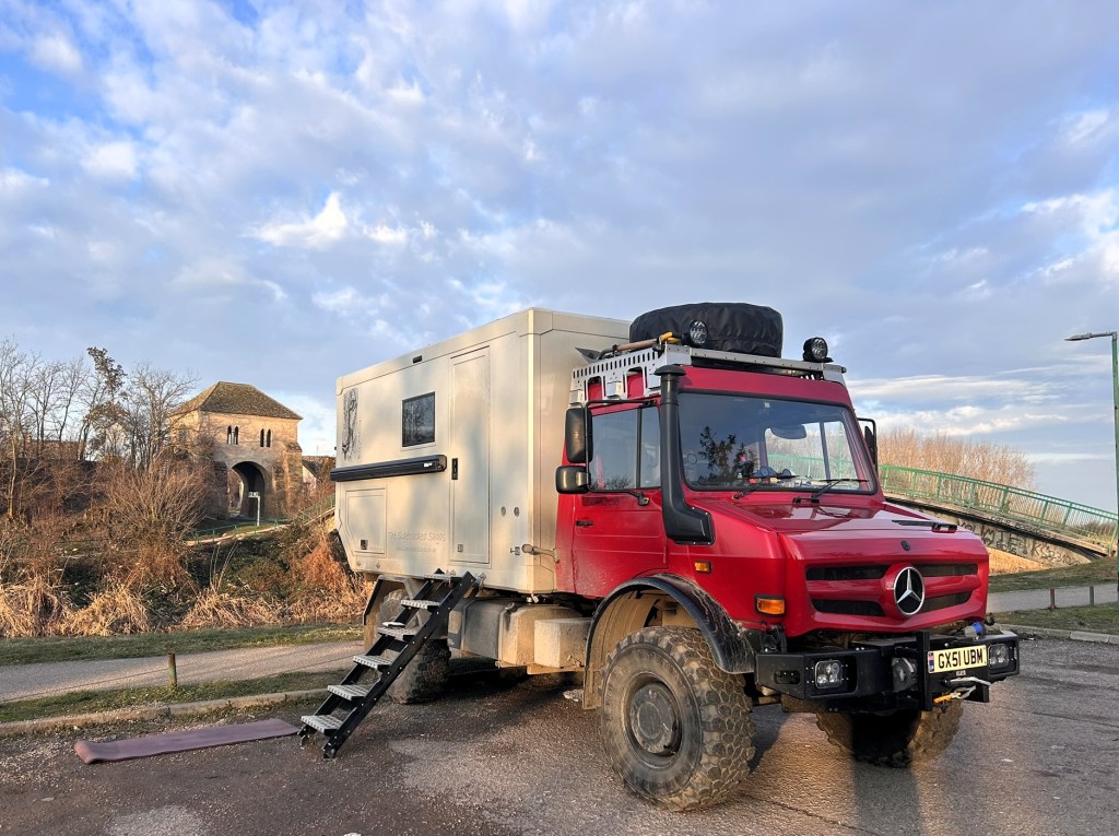 A red Mercedes truck with an attached caravan, parked in a scenic area with a stone structure and bridge in the background. The scene features clear skies and a grassy landscape.