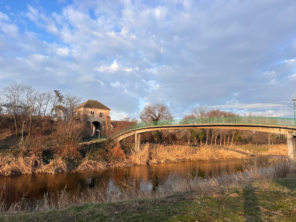 A scenic view of a bridge arching over a river, with a stone structure on the riverbank and trees in the background under a cloudy sky.