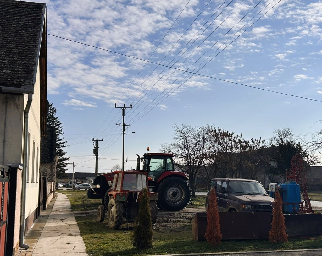 A rural scene featuring two tractors parked next to each other on a grassy area, with a modest house and power lines in the background under a partly cloudy sky.