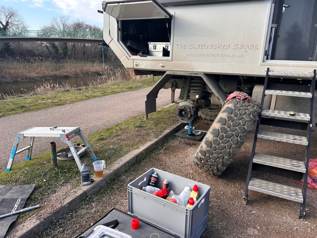 An off-road vehicle with one wheel removed, parked beside a river. Tools and equipment are scattered on the ground, and a folding step ladder is visible.