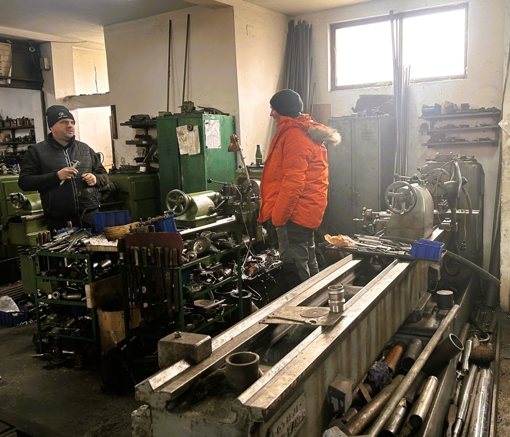 Two men in a workshop surrounded by various tools and machinery, one wearing an orange jacket and the other in a black coat, engaged in conversation.
