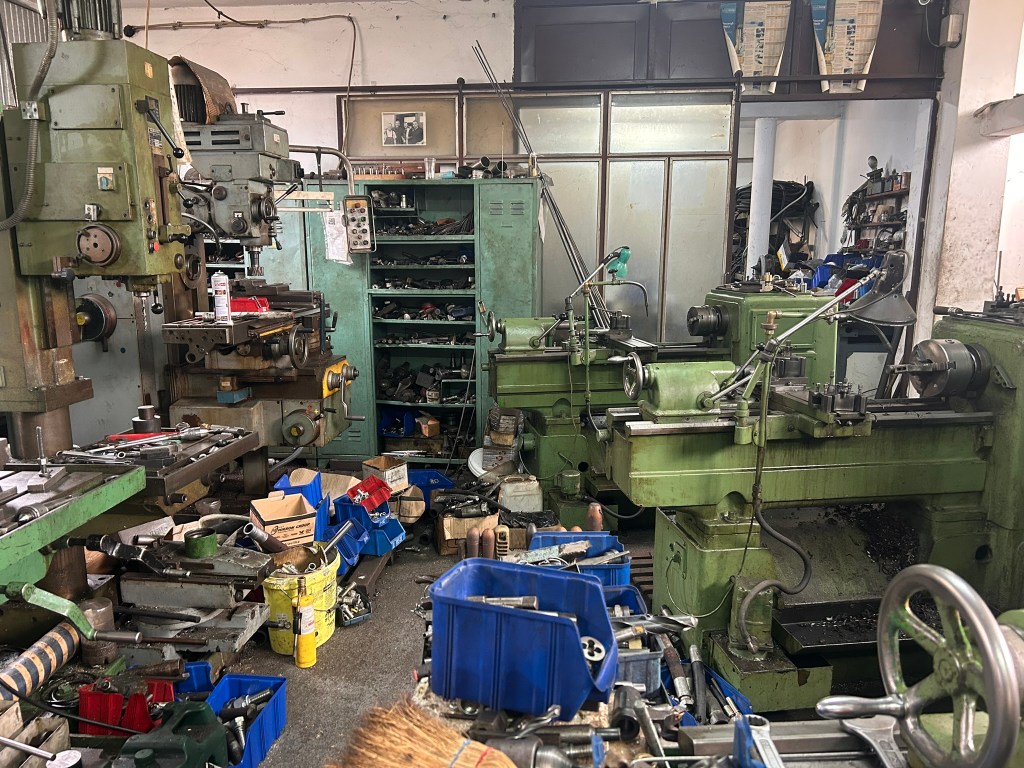 An industrial workshop filled with green machinery, including lathes and milling machines. The area is cluttered with tools and equipment, with toolboxes and parts scattered across the floor.