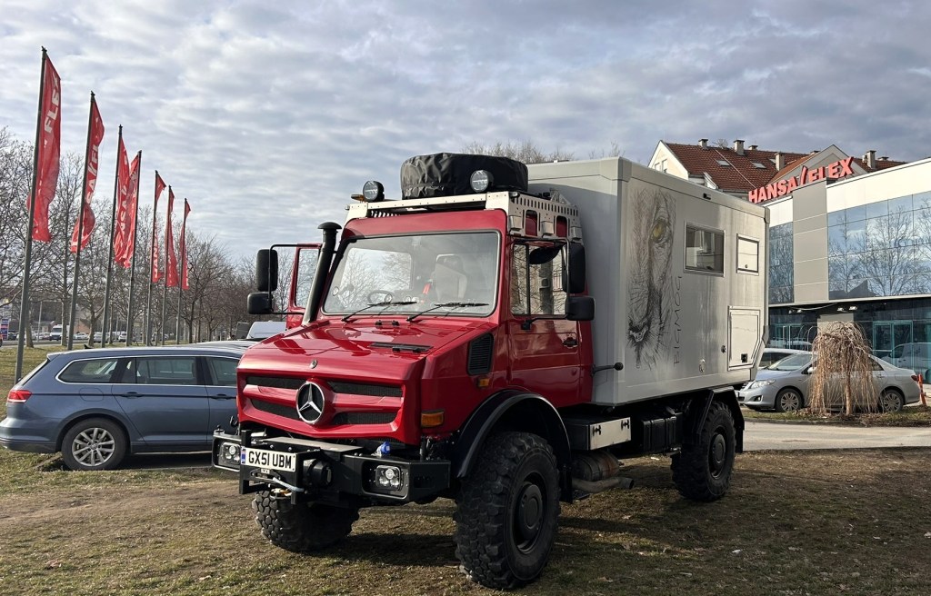 A red Mercedes truck with an attached white camper unit featuring a lion design, parked on grass near buildings and flag poles.