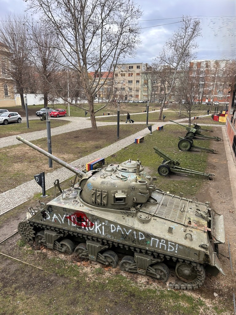 A weathered tank with graffiti, located in a park setting, surrounded by bare trees and a pathway. In the background, residential buildings and military equipment are visible.