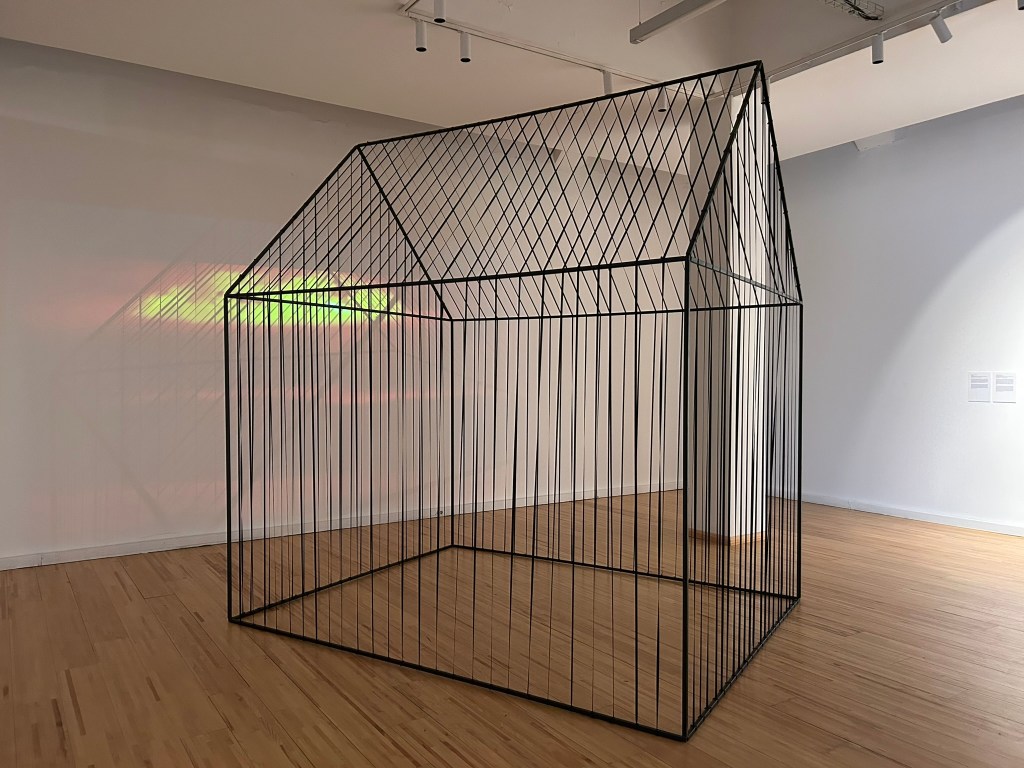 A wireframe sculpture resembling a house, located in an art gallery. The structure features vertical and horizontal lines, creating a geometric appearance with shadows cast on the wall.