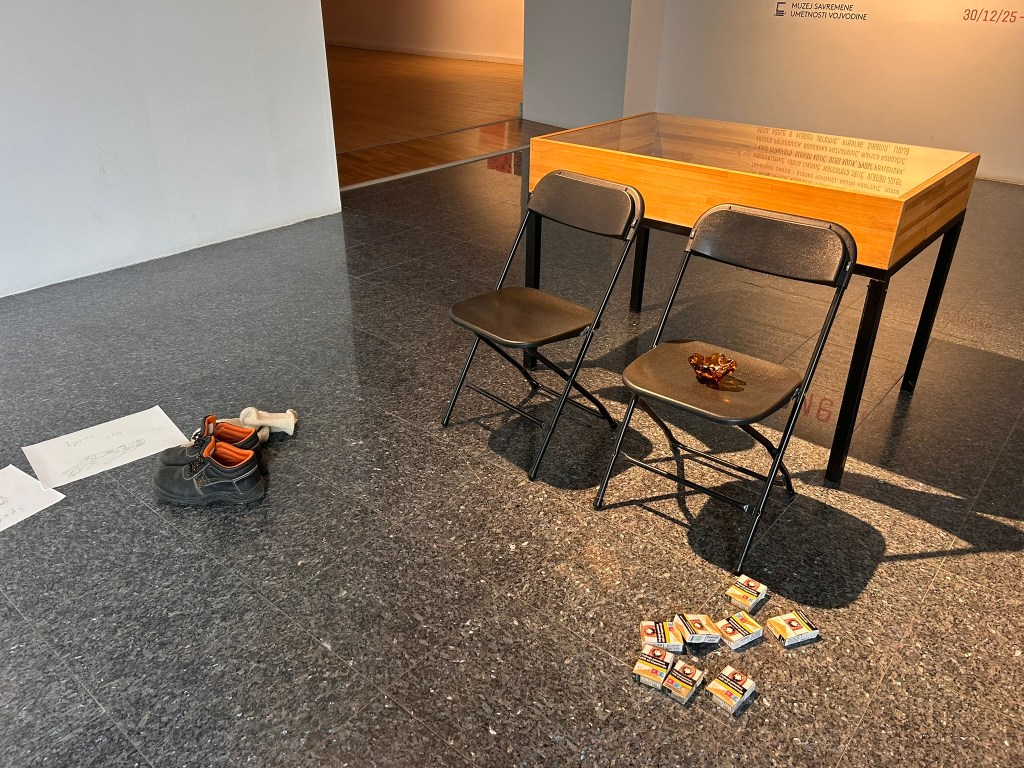 An art installation featuring two black folding chairs and a wooden table, with a small decorative item on one chair, a pair of work boots and a bone on the floor, and several boxes scattered nearby.
