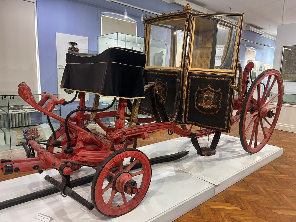 A vintage horse-drawn carriage displayed in a museum, featuring a black canopy and ornate gold detailing on its wooden body, with red wheels and a distinctive emblem.