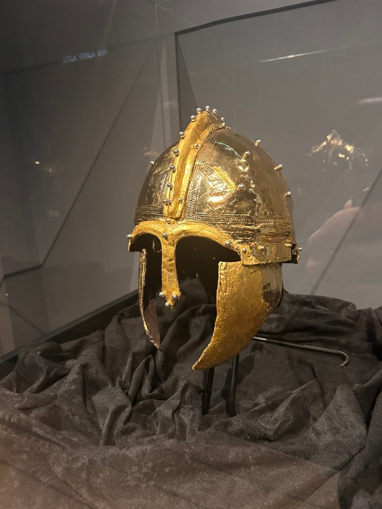 A golden ancient helmet displayed on a stand against a dark background, showcasing intricate designs and detailing.
