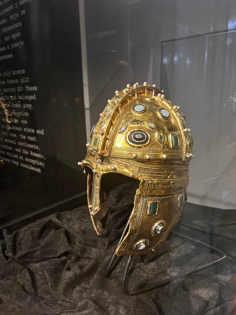 A golden ancient helmet displayed in a museum, intricately decorated with jewels and patterns, resting on a dark fabric background.