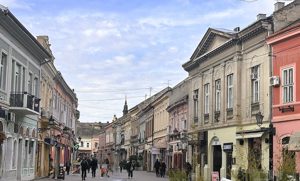 A historic street lined with charming, pastel-coloured buildings, featuring shops and cafés, with people walking along a cobblestone pathway under a blue sky.