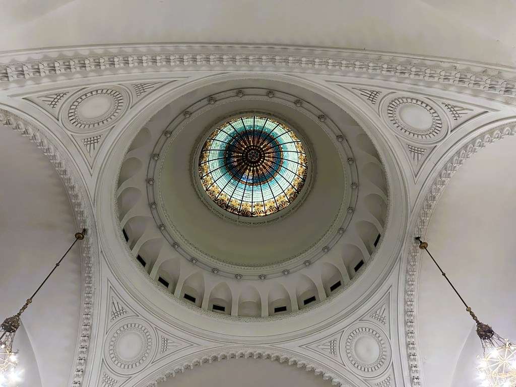 View of a decorative domed ceiling featuring intricate white moulding and a central stained glass window.