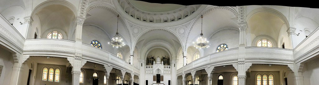 Interior panoramic view of a grand hall with high, ornate ceilings, decorative mouldings, and stained glass windows, featuring chandeliers and a balcony.