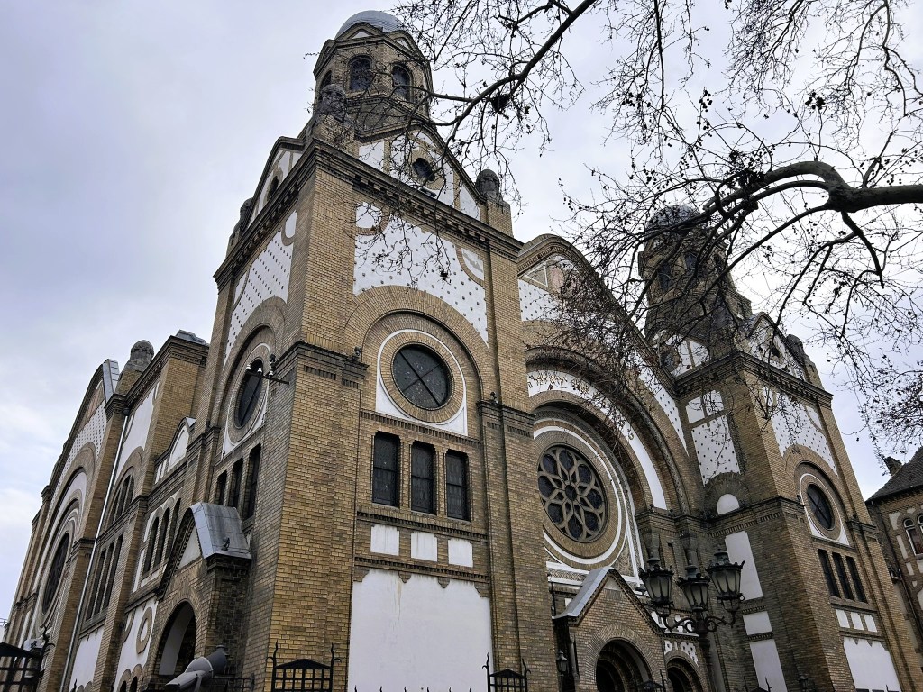 A close-up view of an ornate brick building with large windows and a prominent dome, surrounded by bare trees and a cloudy sky.